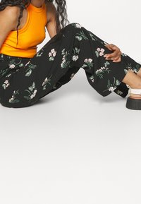 Femme assise, portant un haut orange sans manches, un pantalon noir avec un imprimé floral vert et rose, et des sandales à plateforme blanches.