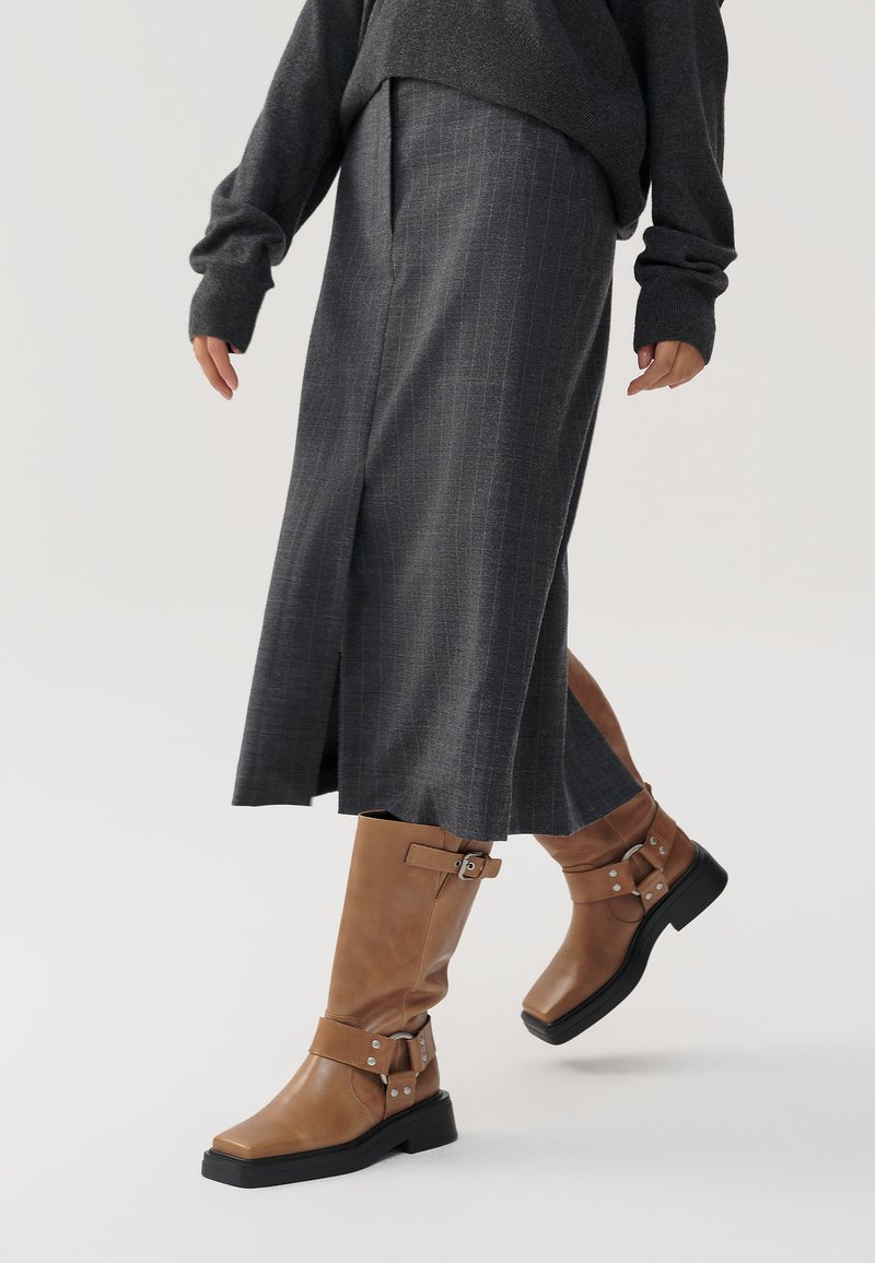 Botas de cuero marrón hasta la rodilla con punteras cuadradas, detalles de hebilla y una suela negra gruesa, combinadas con una falda midi de rayas grises.