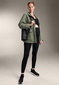 Tűzött zöld dzseki, fekete crop top, fekete leggings és fekete sportcipő. Hordoz egy pántos, feltekert jóga matrat. Egyszerű, praktikus dizájn.