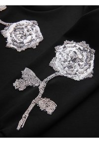 Motifs de roses argentées perlées et pailletées sur tissu noir, montrant la fleur, la tige et les feuilles en gros plan détaillé.