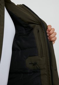 Interior de chaqueta verde oliva con forro negro, con un bolsillo con cremallera y un detalle de cierre ajustado con cordón. Estilo acolchado texturizado.