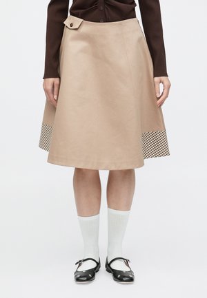Ein beiger A-Linien-Rock mit seitlicher Knopfleiste und kariertem Muster am Saum. Die Materialien wirken glatt mit einem strukturierten Design.