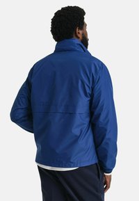 Chaqueta de nylon azul, ligera, con un cuello alto, puños elásticos y una costura horizontal en la espalda. Textura suave con un acabado mate.