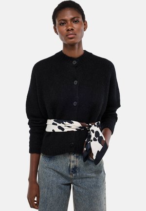 Femme portant un cardigan noir boutonné avec une ceinture-écharpe à motifs blancs et noirs, assorti à un jean bleu clair, sur un fond uni.
