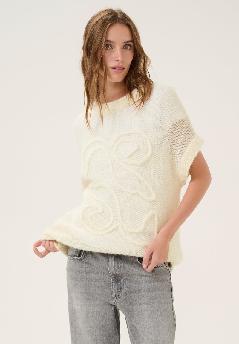 Knit jumper in soft cream with a textured, raised abstract design. Short sleeves and a loose fit; worn over gray jeans.