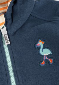 Navy-Fleecejacke mit einem türkisfarbenen Reißverschluss, gestreiftem Kapuzenfutter und einem bunten Flamingo-Patch mit Rollschuhen.