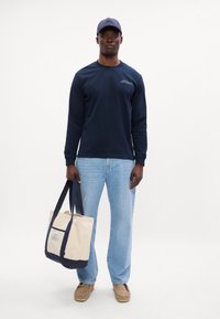 Mere pikkade varrukatega navy särk, heledad sinised teksad, beežid kingad, hoiab käes beeži kott, millel on tume sinine põhja ja silt, ühtlane taust.