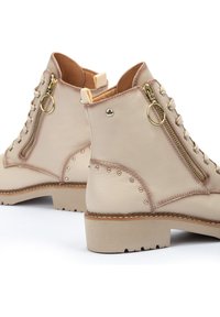 Botines de cuero beige con herrajes dorados, cordones en la parte frontal, cremallera lateral y acentos de tachuelas. Suela resistente con un ligero acabado en el tacón.