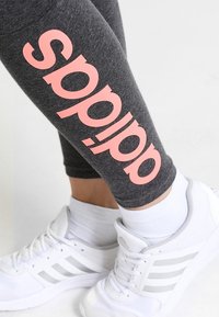 Szare legginsy z dużym różowym logo "sp", w połączeniu z białymi butami sportowymi z szarymi akcentami i trzema paskami. Tekstura jest gładka.