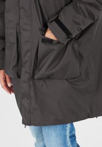 Veste noire isolée avec un col haut, fermeture velcro visible et grandes poches avant. Le tissu semble lisse et résistant à l'eau.
