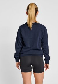 Sudadera azul marino de corte relajado, con puños y dobladillo acanalados. Se lleva con pantalones cortos negros ajustados, que muestran una textura suave y costuras mínimas.