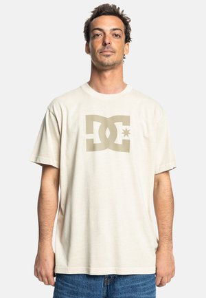 Beige Baumwoll-T-Shirt mit einem großen olivgrünen DC-Logo und Stern-Grafik auf der Vorderseite. Lockerer Schnitt und kurze Ärmel. Glatte Textur.