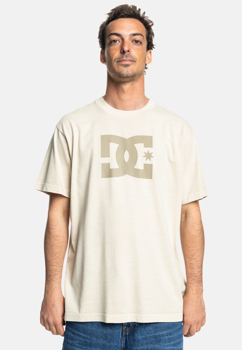 Beige bavlněné tričko s velkým olivově zeleným logem DC a hvězdou na přední straně. Volný střih a krátké rukávy. Hladká textura.