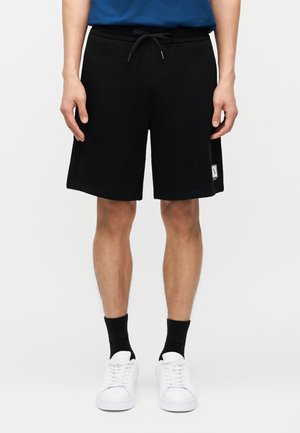 Άνδρας που φοράει μαύρα σορτς μέχρι το γόνατο με κορδόνι, μαύρες κάλτσες, λευκά αθλητικά παπούτσια και μπλε πουκάμισο σε απλό λευκό φόντο.