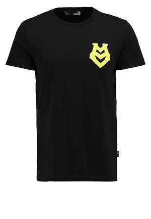 Homme portant un t-shirt noir avec un logo jaune sur la poitrine, un pantalon sombre et des baskets blanches avec des accents verts, debout devant un fond blanc.