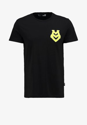 Man draagt een zwart T-shirt met een geel logo op de borst, donkere broek en witte sneakers met groene accenten, staand tegen een witte achtergrond.