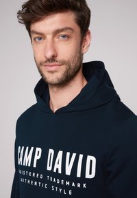 Granatowy hoodie wykonany z miękkiego materiału, z kangurzą kieszenią i odważnym białym napisem "CAMP DAVID" na klatce piersiowej.