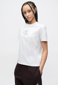 Tricou alb din bumbac cu mâneci scurte, cu logo-ul „CK” cusut în gri deschis pe piept. Textură netedă, decolteu clasic rotund.