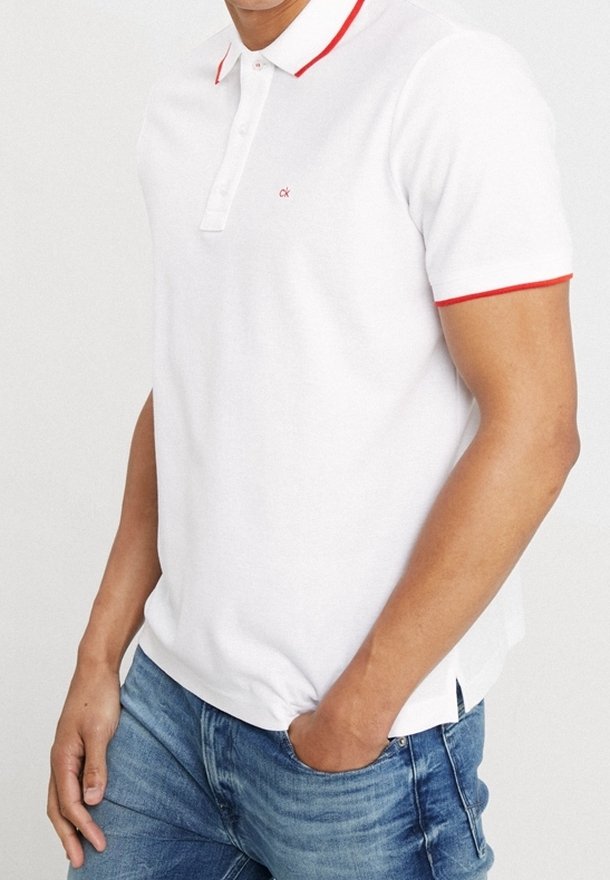 Homme portant un polo blanc à manches courtes avec un liseré rouge sur le col et les manches, associé à un jean bleu, main dans la poche.