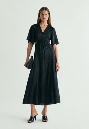 Mujer con un vestido midi negro con cortes en la cintura, sosteniendo un clutch negro y llevando mules negras con tacón y pendientes de aro plateados.