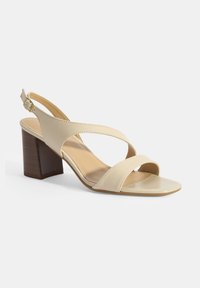 Sandale à talon haut en cuir beige avec un bout ouvert, une sangle à l'arrière et des bandes croisées à l'avant. Talon en bloc avec une texture semblable à celle du bois.