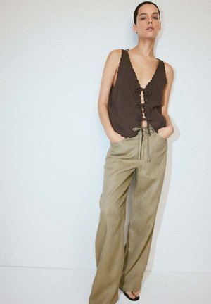 Mujer con una blusa sin mangas marrón oscuro con nudo delantero y pantalones beige holgados, de pie con las manos en los bolsillos.