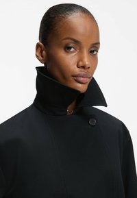 Czarny trencz z wysokim kołnierzem, zapinany na guziki, wykonany z fakturowanej tkaniny. Projekt ma elegancką, dopasowaną sylwetkę.