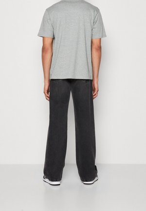 Mand iført en lysegrå kortærmet t-shirt, mørke vide jeans og sorte og hvide sneakers, stående med ryggen til på en ensfarvet baggrund.