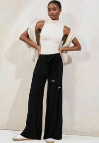 Schwarze weite Hose mit einem gebundenen Bund, kombiniert mit einem ärmellosen beigen Top. Die Accessoires umfassen Creolen und einen leichten Pullover, der über die Schultern gelegt ist.