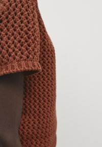 Top lavorato a maglia di colore ruggine con un motivo a trama aperta testurizzato e una sezione solida marrone. I bordi sono arrotolati, migliorando il design.