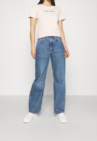 Camiseta de algodón color rosa claro con un logotipo rojo y negro centrado en el pecho, combinada con jeans de pierna ancha de mezclilla azul de cintura alta y zapatillas blancas.