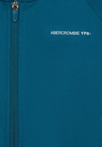 Teal zip-up jacket made of smooth fabric; features a front zipper and white logo text "ABERCROMBIE YPB" on the upper left.