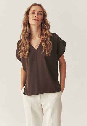 Frau mit langen, welligen blonden Haaren, die eine dunkelbraune V-Ausschnitt-Bluse mit kurzen Ärmeln und weiße Hosen trägt, steht mit den Händen in den Taschen.