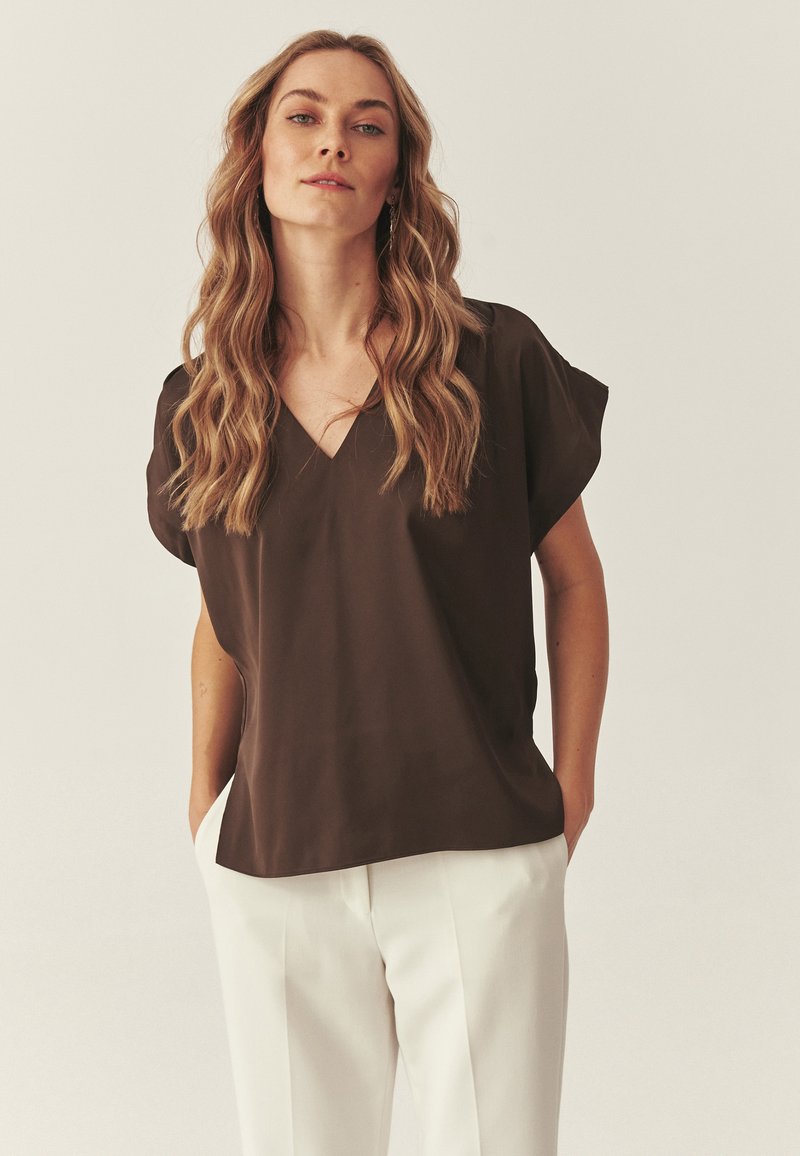 Femme aux longs cheveux blonds ondulés portant un chemisier à manches courtes col en V de couleur marron foncé et un pantalon blanc, debout les mains dans les poches.