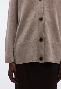 Helepruun kardigan pehmeast kootud materjalist, millel on ees tumedate pruunide nuppudega kinnitused ning soonilised mansetid ja alumine äär.