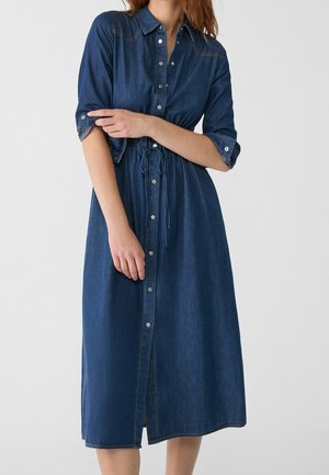 Femme portant une robe en jean mi-longue boutonnée avec des manches retroussées et une taille à cordon, debout devant un fond uni.