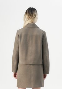 Žena s dlouhými blond vlasy, která má na sobě odpovídající taupe sametovou bundu a sukni, stojí zády k fotoaparátu na bílém pozadí.