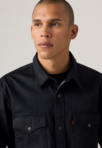 Dark denim shirt featuring a pointed collar, two chest pockets with button closures, and subtle stitching details. Smooth texture.