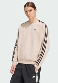 Svetlobéžová mikina s čiernymi pruhmi na rukávoch, okrúhlym výstrihom a malým logom na hrudi. Mäkká látka, voľný strih, rebrované manžety.