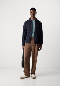 Chaqueta azul marino de botones sobre una camisa verde claro, combinada con pantalones de pierna ancha marrones y zapatillas blancas con detalles en marrón; sosteniendo un bolso negro.