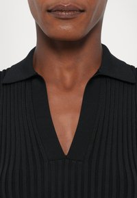 Fekete bordázott kötött felső V-nyakkal és gallérral, sima textúrával és testhez simuló dizájnnal. Nagyítás a nyakrészre.