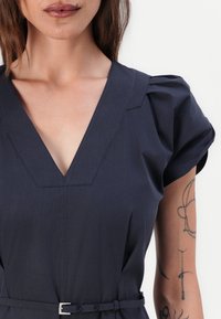 Dunkelblaues Kleid mit V-Ausschnitt, kurzen Puffärmeln und tailliertem Gürtel. Weicher Stoff mit subtilen Stickdetails und minimalistischer Gestaltung.