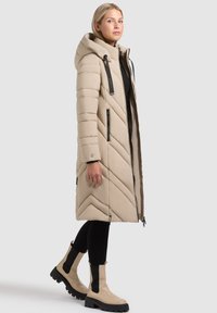 Bež puhasta bunda s kapuco, s šivanim vzorcem v obliki cik-cak, črnimi poudarki, dvojno sprednjo zadrgo in ohlapno silhueto.