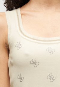 Gros plan d'une personne portant un débardeur en tricot crème avec de petits motifs symétriques en strass sur tout le tissu.