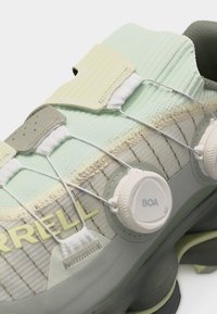 Gros plan sur une chaussure de sport vert clair et grise avec un système de laçage à molette BOA et le logo "Merrell" sur le côté.