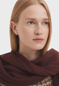 Écharpe bordeaux en matériau doux, drapée autour du cou, avec une apparence légère et texturée, complémentant un pull à motifs.