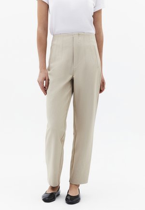 Close-up van een persoon die beige, hoogwaterbroeken, een wit shirt met korte mouwen en zwarte ballerina's draagt tegen een witte achtergrond.