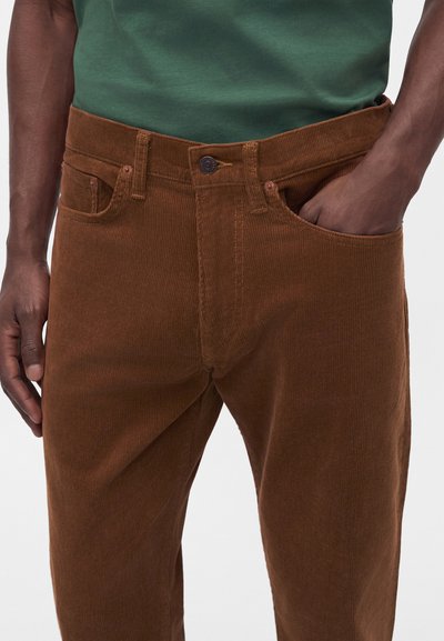 Uomo che indossa pantaloni di velluto a coste marroni con una mano in tasca e camicia verde visibile all'altezza della vita.