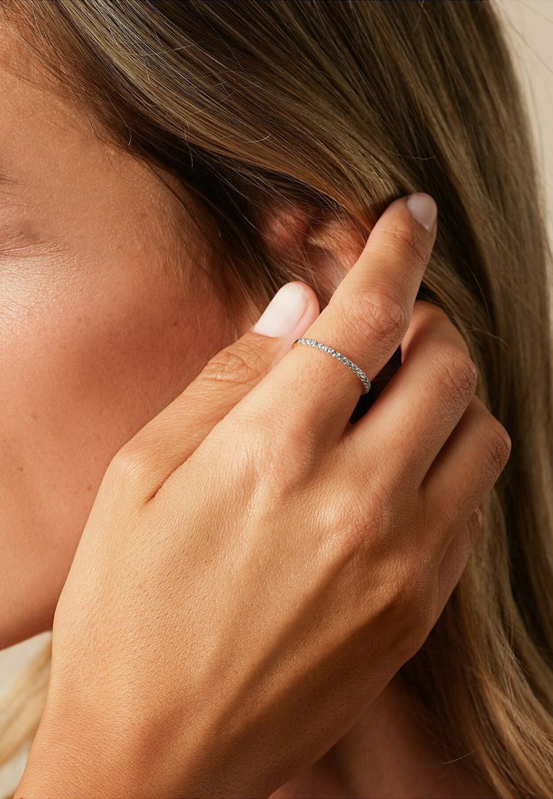 Silver ring med små, klara stenar, bärs på en hand som rör vid hår. Hudtonen är varm med en slät yta. Korta naglar, naturlig färg.