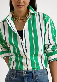 Πουκάμισο με πράσινες και λευκές ρίγες και μπλε γραμμές, με γιακά και μανίκια που είναι γυρισμένα. Φορεμένο πάνω από σκούρο τοπ και τζιν παντελόνι.